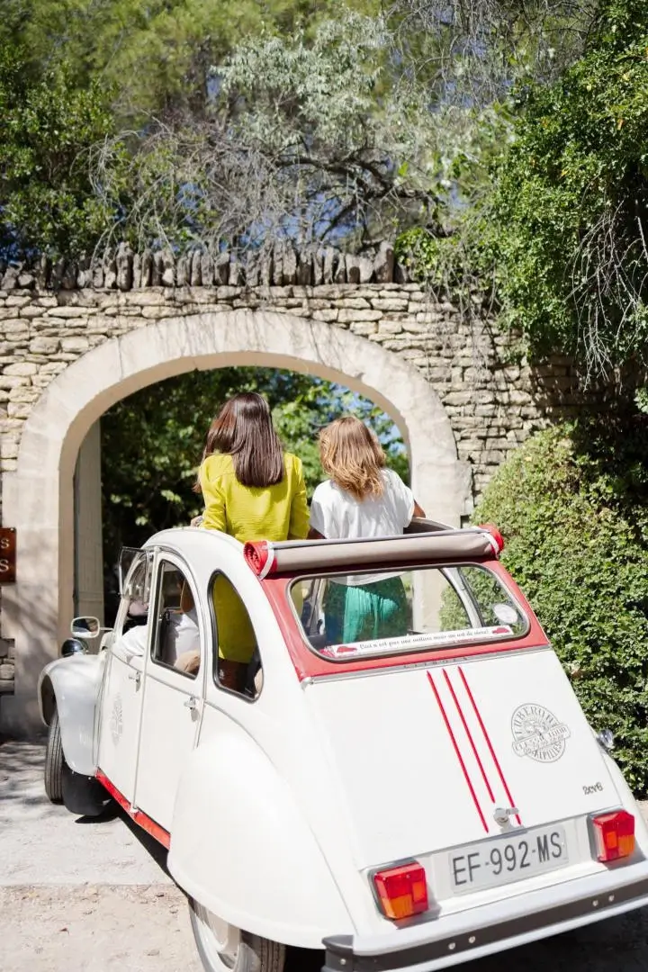2CV blanche à bandes rouges, deux passagers debout par le toit ouvert, devant une arche en pierre