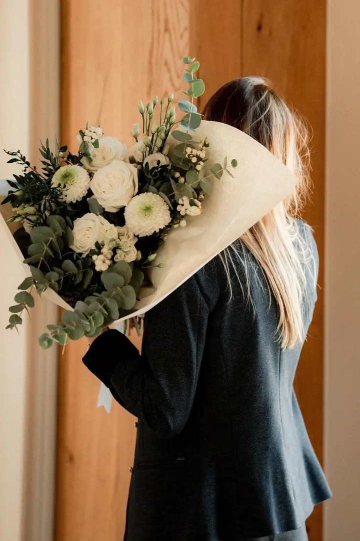 Femme de dos portant un grand bouquet de fleurs blanches et de feuillage emballé dans du papier